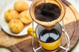 cafe-sendo-coado-no-coador-com-uma-caneca-amarela-abaixo-com-um-prato-de-pao-de-queijo-ao-fundo